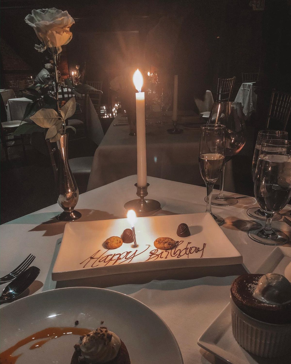 Birthday dessert plate with chocolate writing, a lit candle, and champagne in a restaurant.