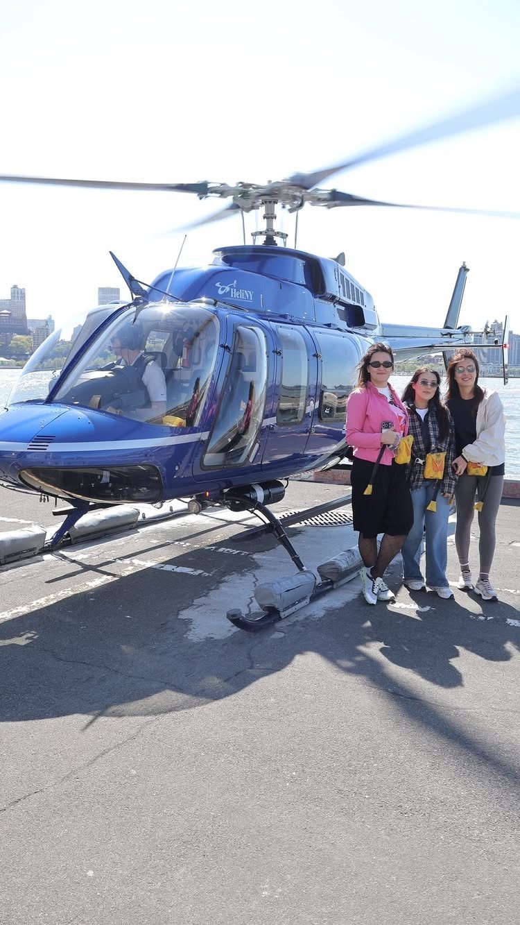 Three women posing next to a blue helicopter on a sunny landing pad.