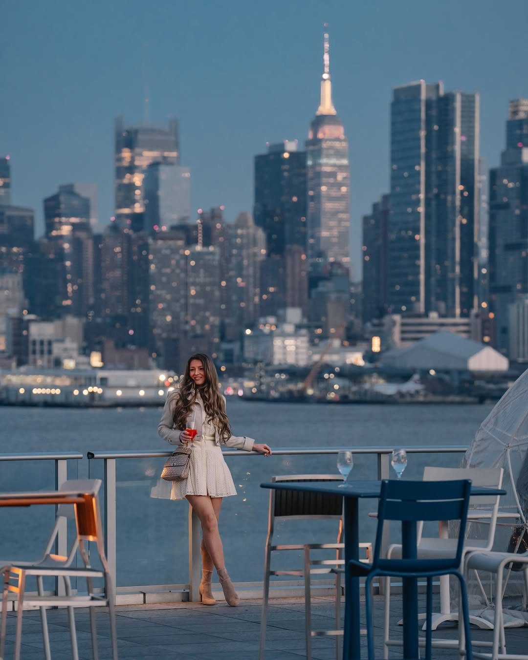 Woman in white dress holding a drink on a rooftop with NYC skyline background