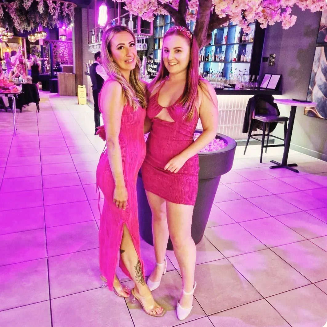 Two women in pink dresses posing in a venue with pink lighting.