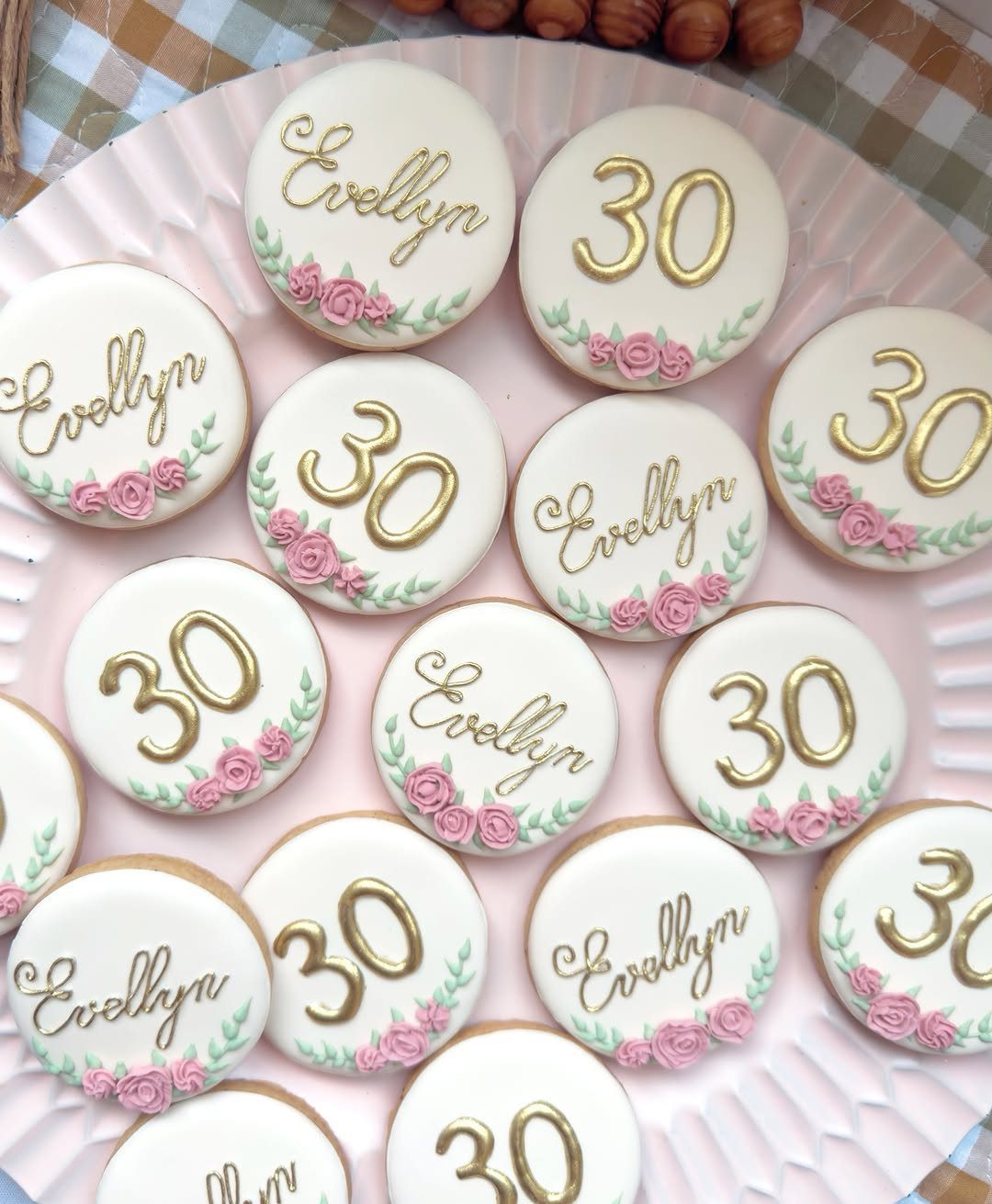 Round white iced cookies with gold numbers and names decorated with pink roses.