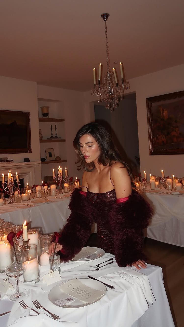 Woman in red fur wrap standing by a candlelit dinner table with white linens.
