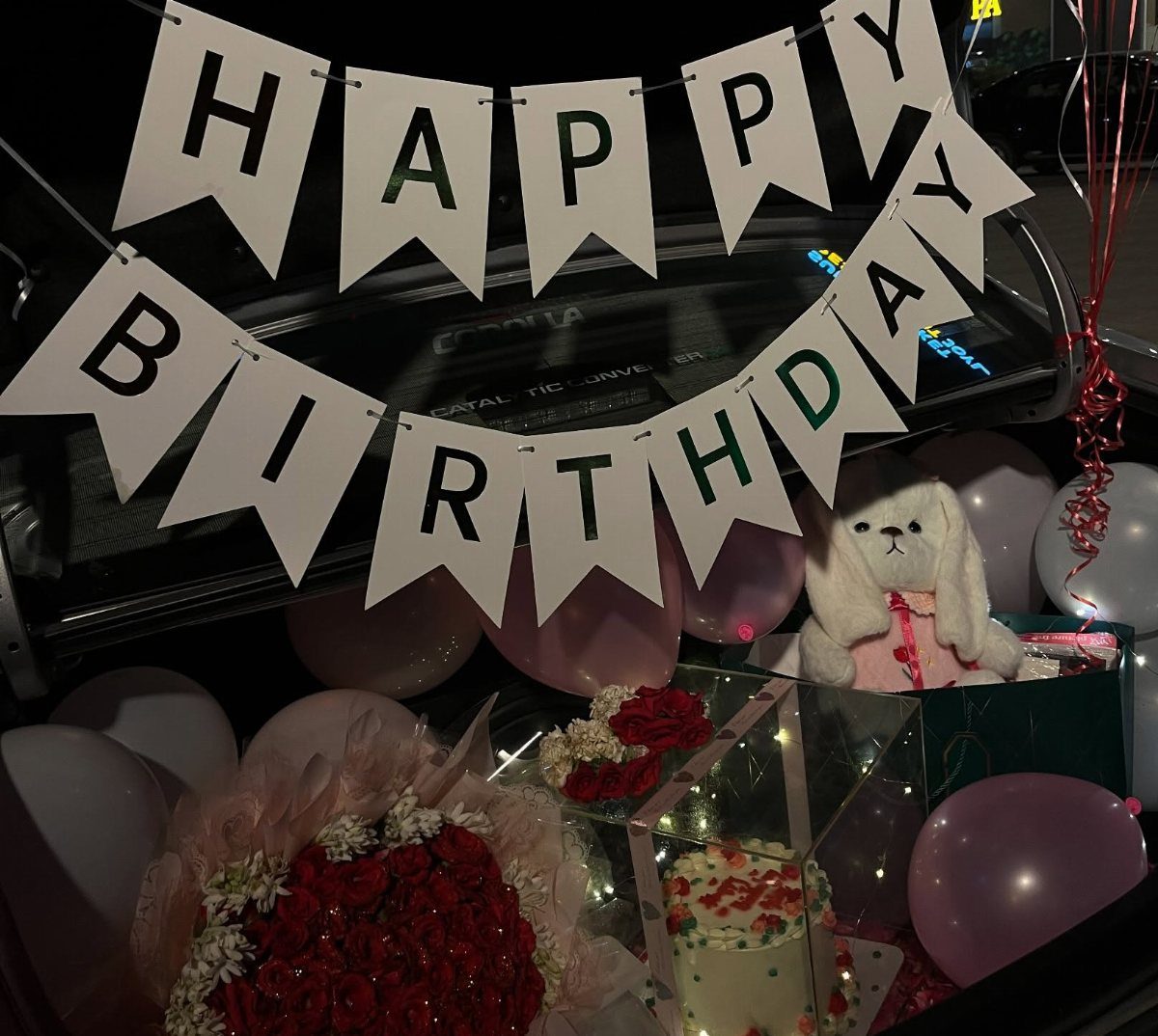Car trunk decorated with birthday banner, balloons, roses, cake, and a stuffed bunny.