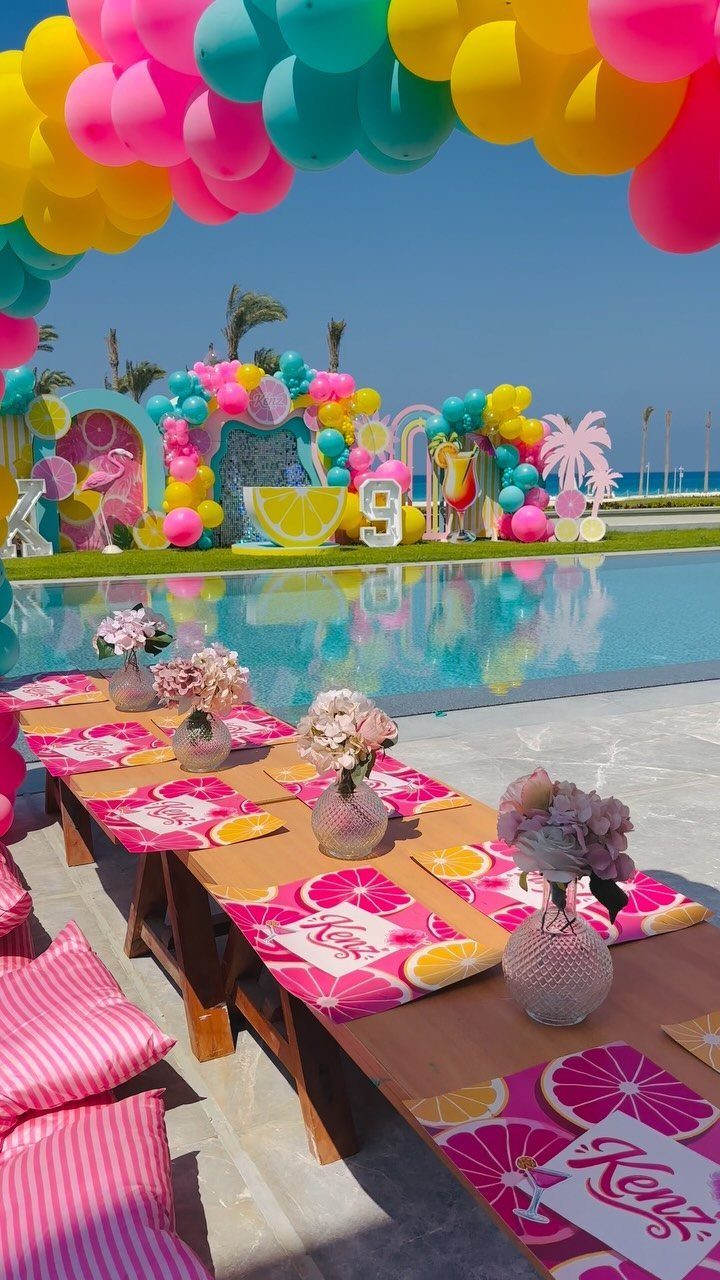 Poolside party setup with colorful balloon arch, wooden table, and pink grapefruit placemats.