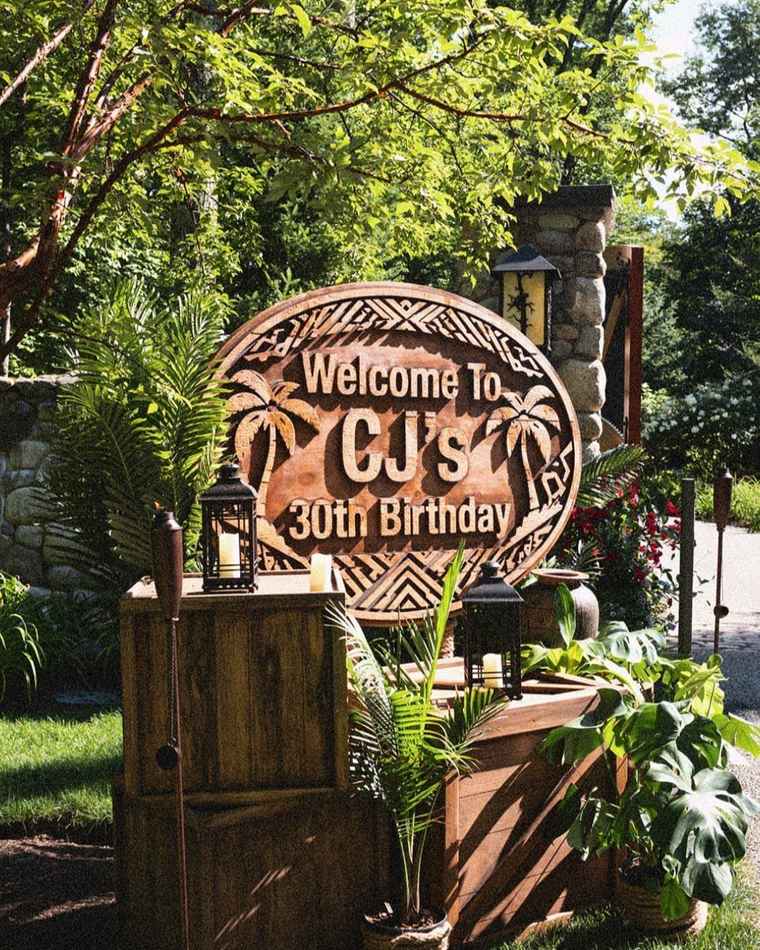 Wooden tropical welcome sign for CJ's 30th birthday surrounded by lush green plants.