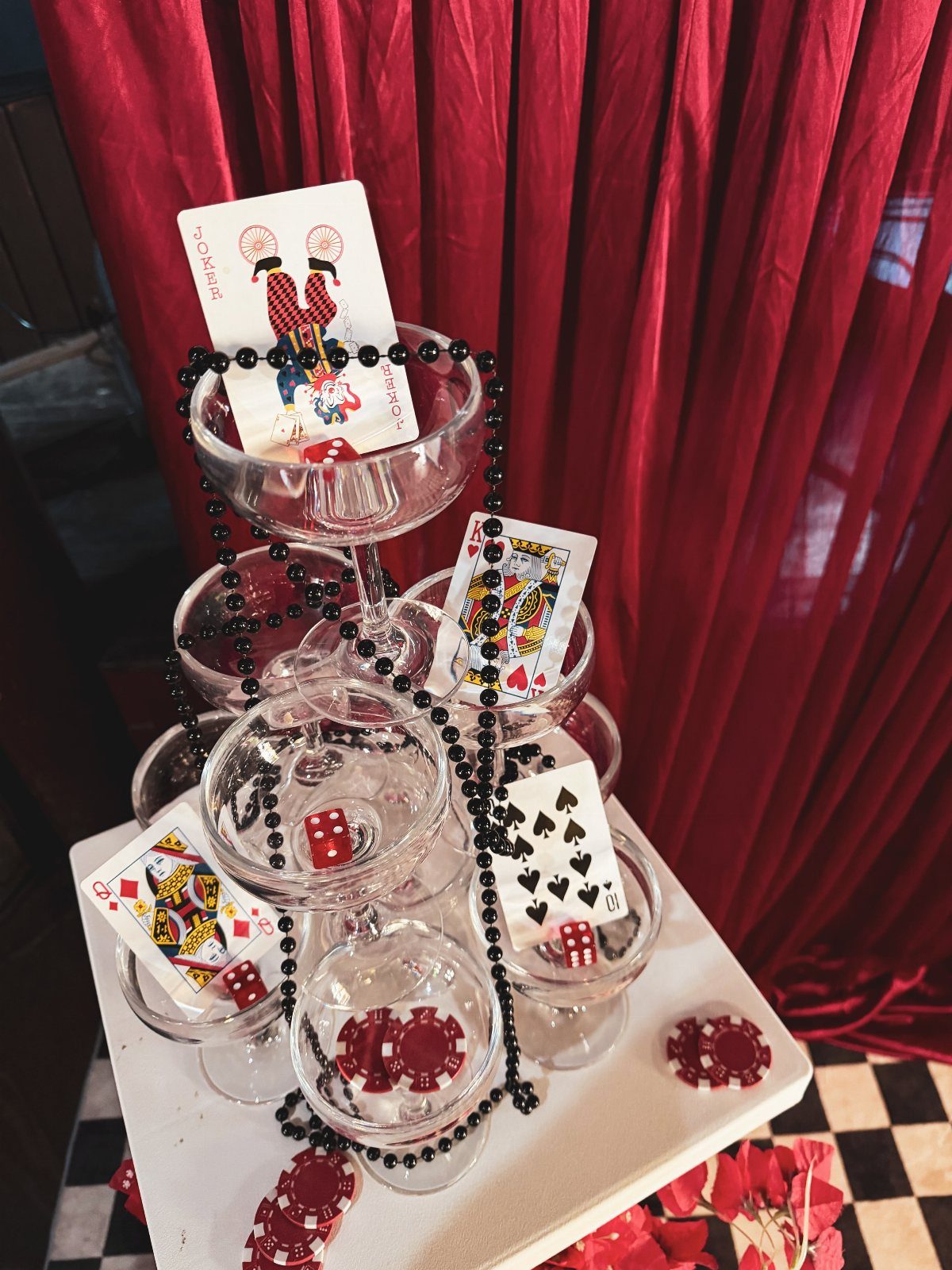 Champagne glass tower decorated with playing cards, dice, and poker chips against red curtains.