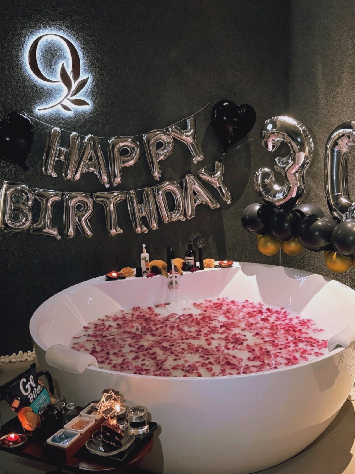 White bathtub with pink petals and silver 30th birthday balloons in a room.