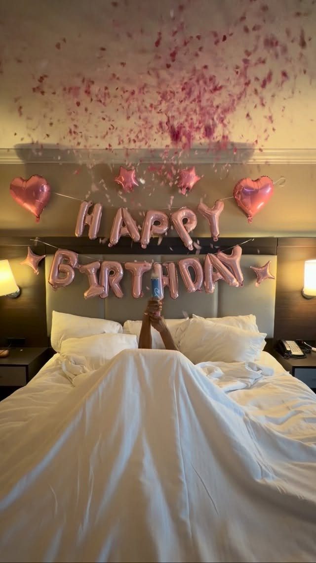 Person in a hotel bed with pink birthday balloons and foil lettering above them.