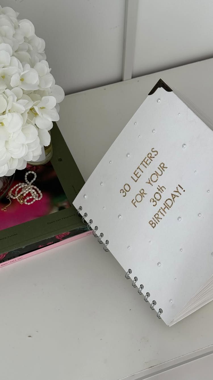 White spiral notebook with gold text for a 30th birthday next to flowers.