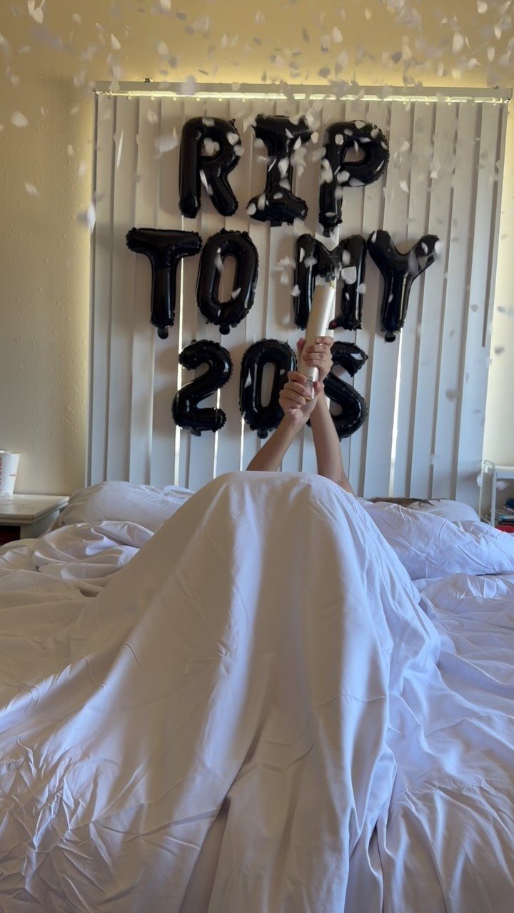 Person under white sheets with black balloons spelling RIP TO MY 20s