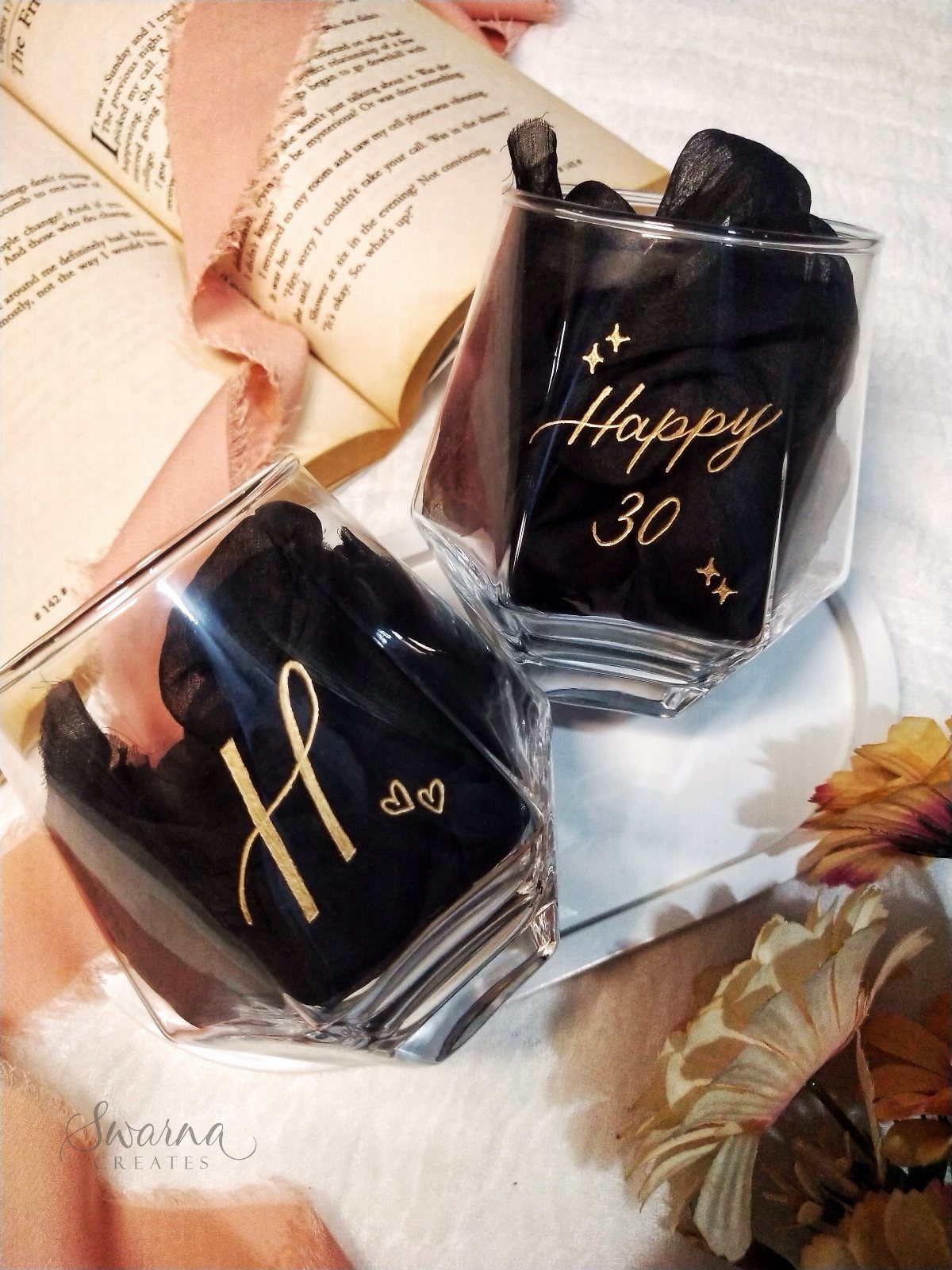 Two personalized glass tumblers with gold lettering and black fabric inside near a book.