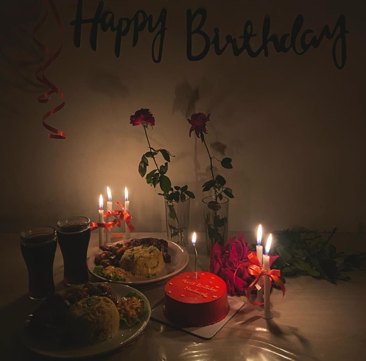 Birthday dinner table with red cake, lit candles, roses, and plates of food.