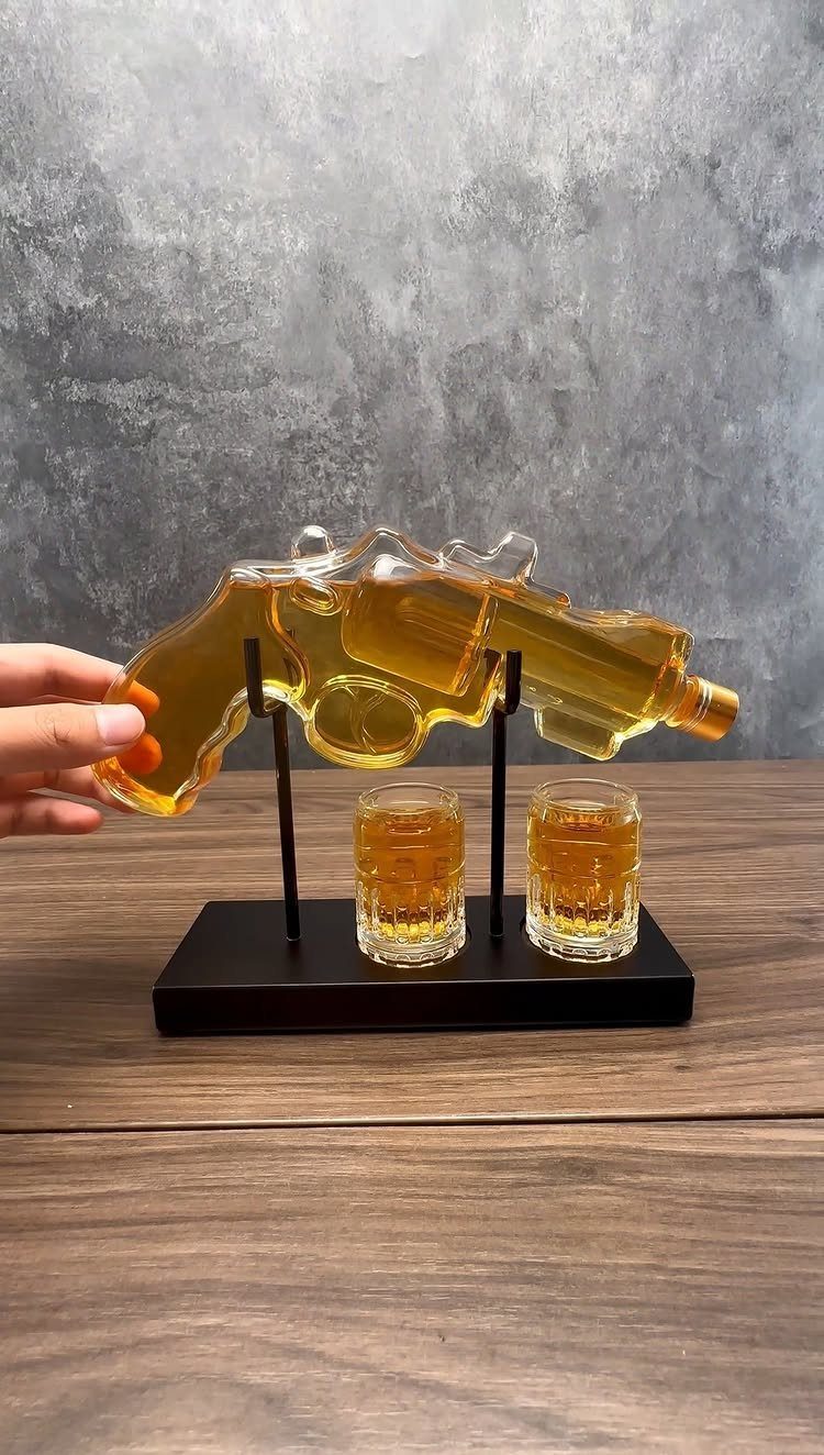 Gun-shaped glass decanter with two shot glasses on a black stand.