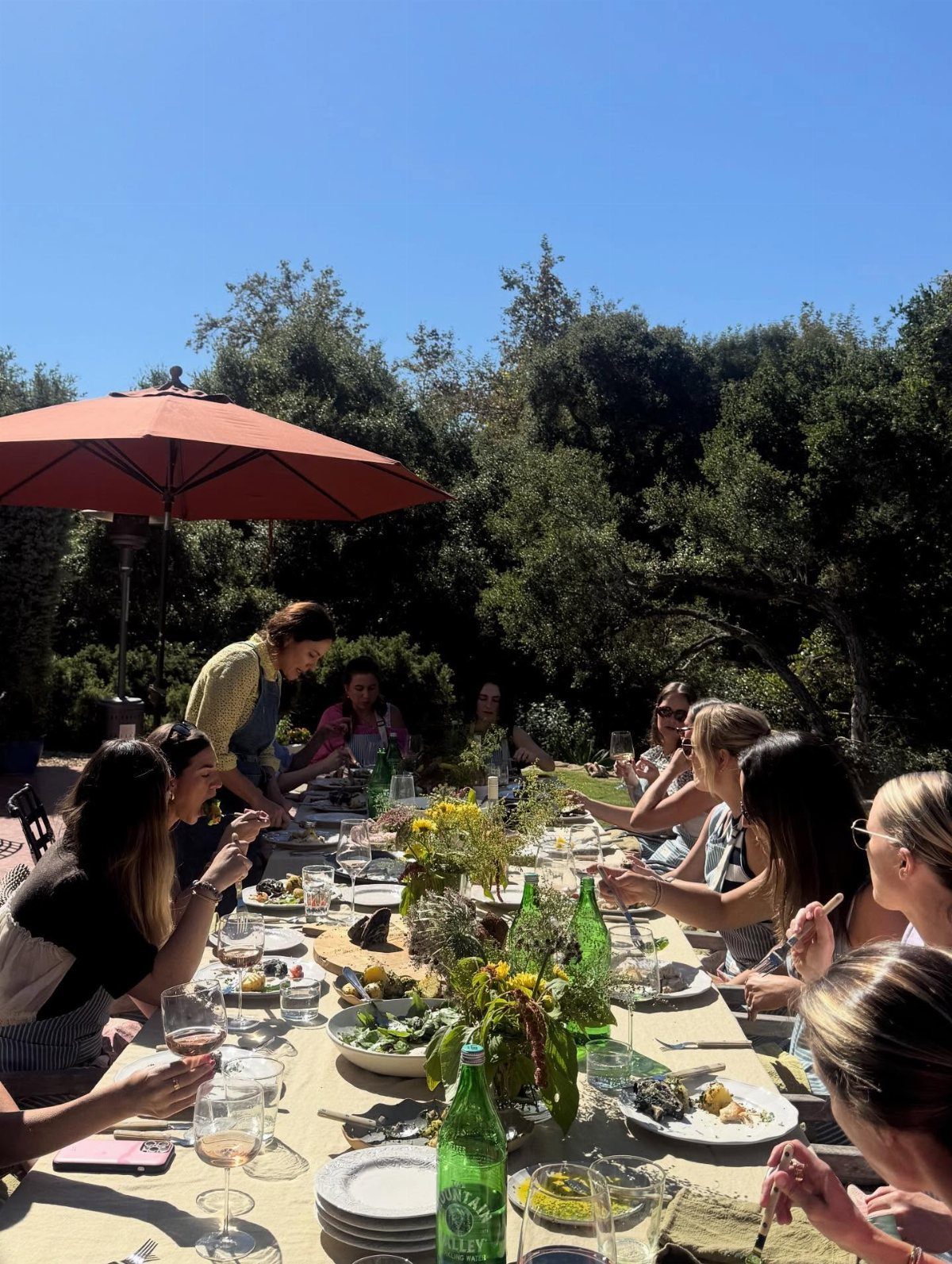 Women enjoying a meal together at a long outdoor table with floral decorations.