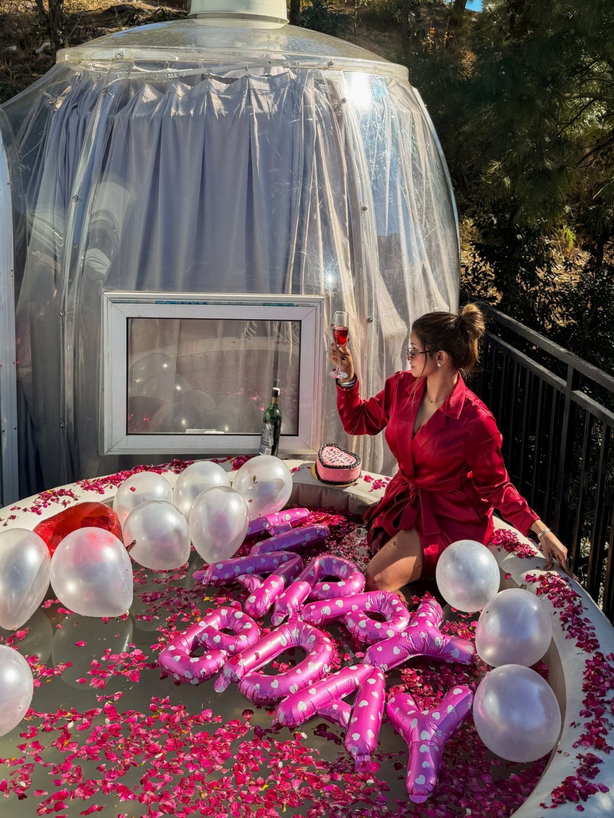 Woman in red robe celebrating birthday with balloons and petals by a glamping dome.
