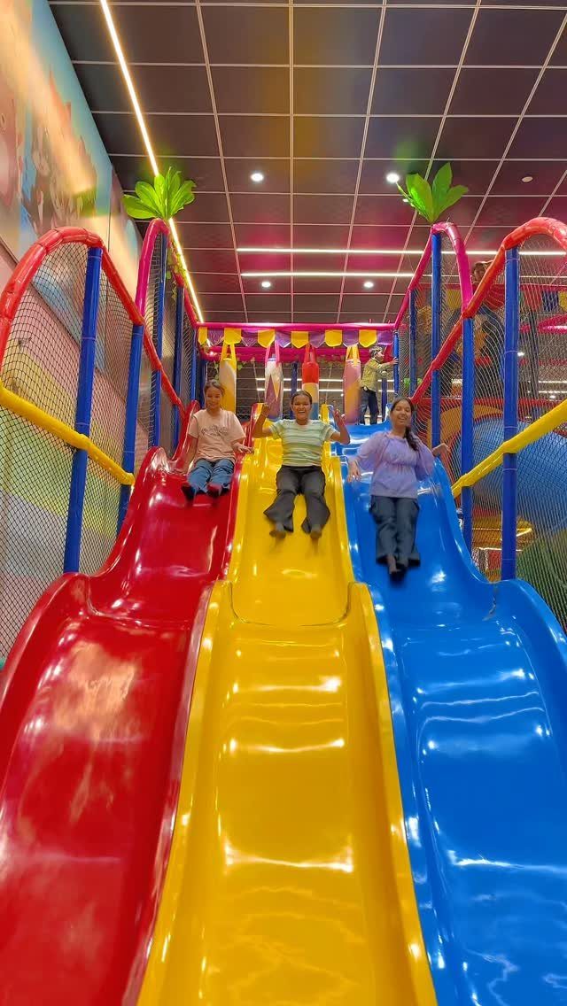 Three women sliding down colorful red, yellow, and blue slides in a play center.