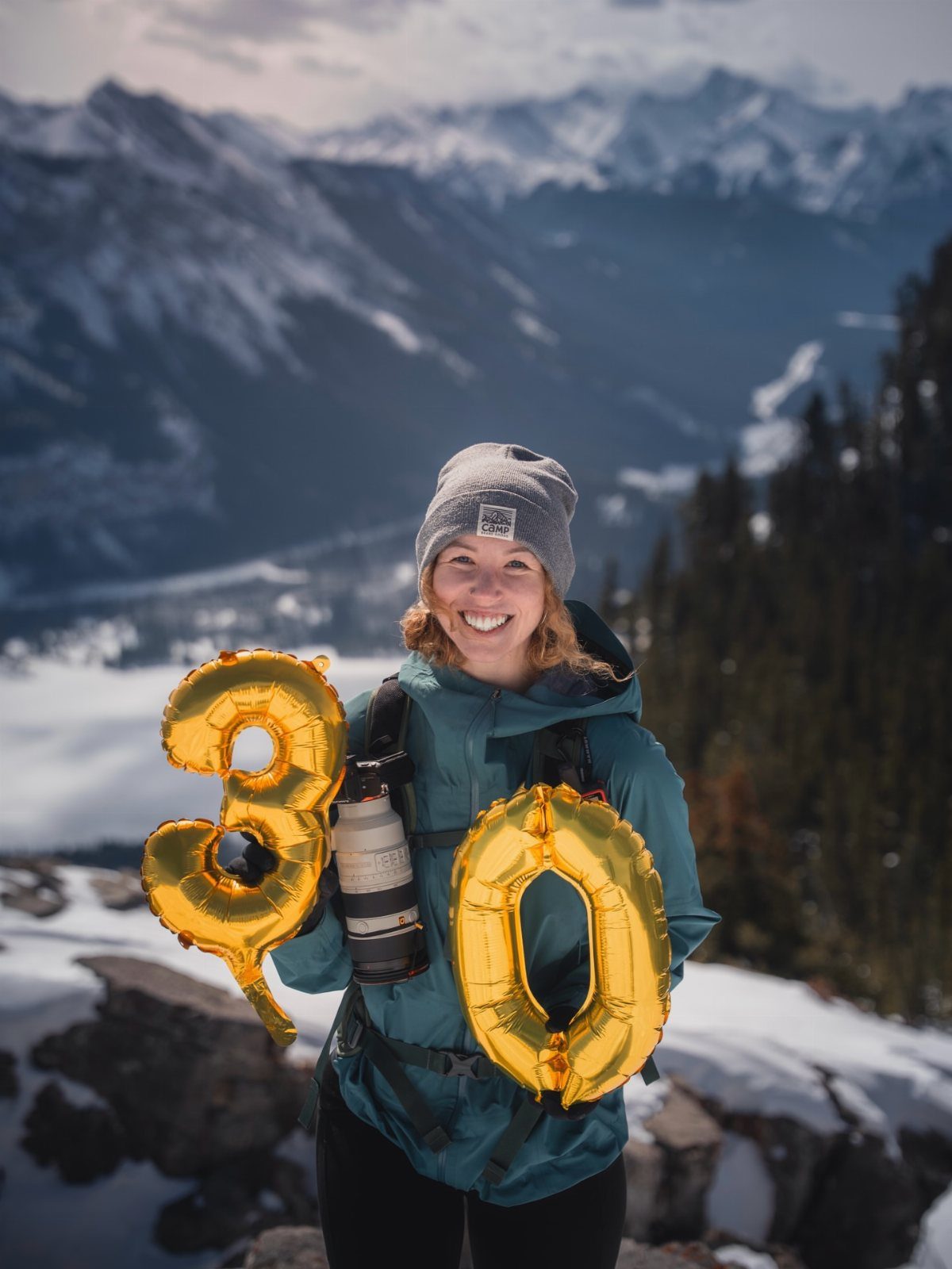 Woman in hiking gear holding gold 30 balloons on a snowy mountain peak