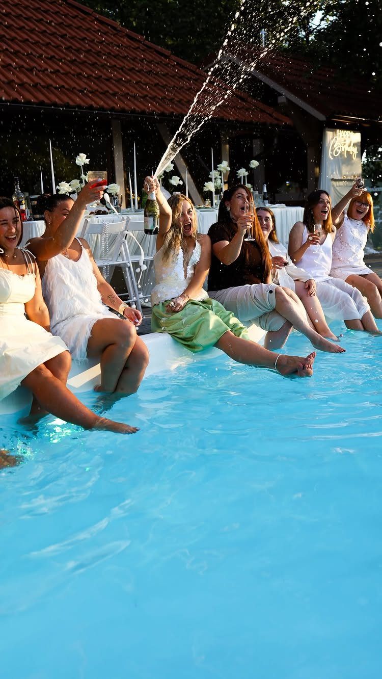 Women in white dresses celebrating by a pool with popping champagne spray.