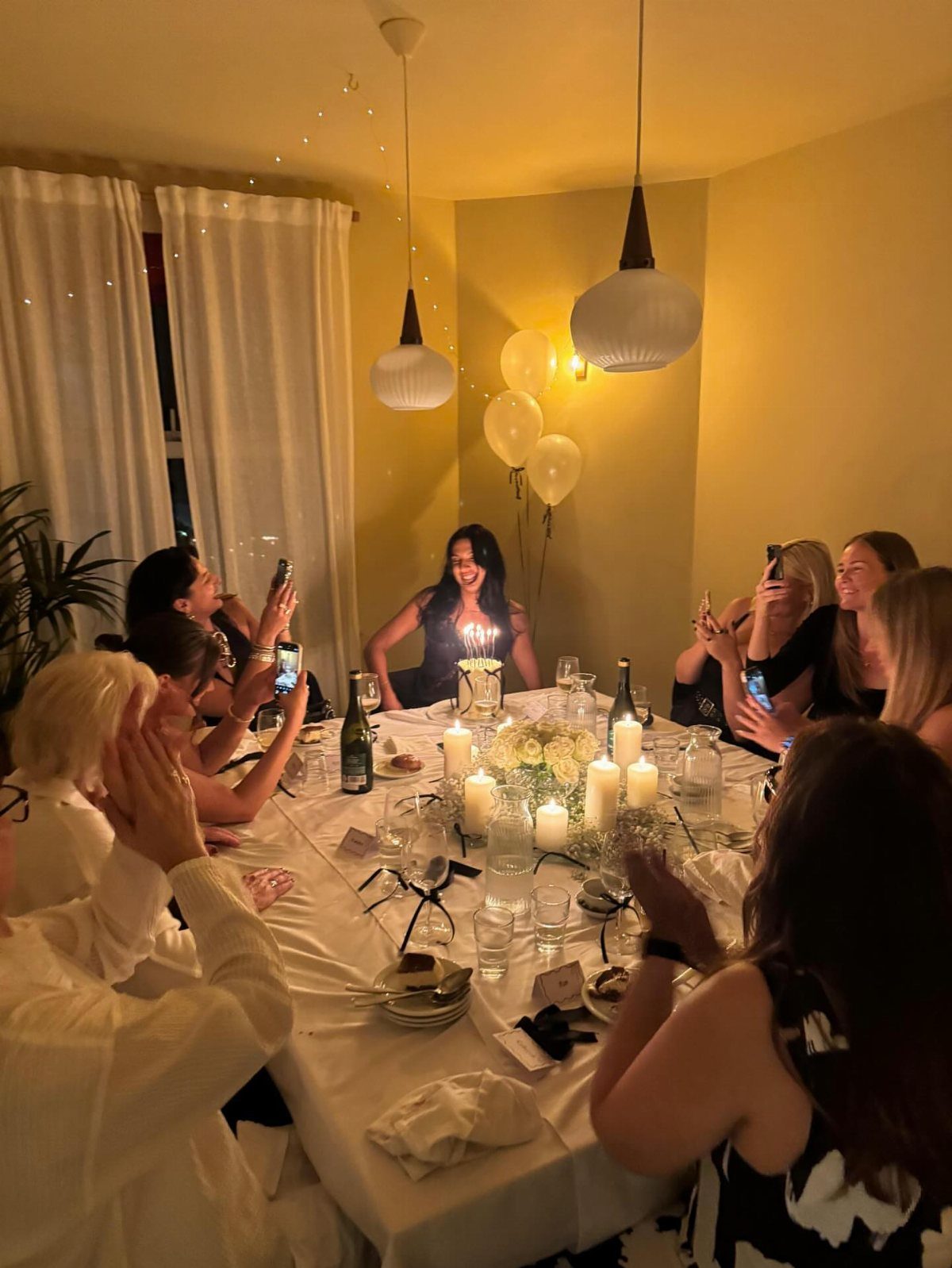 Women celebrating a birthday around a candlelit dinner table with white balloons.