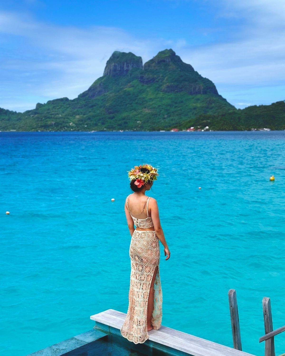 Woman in gold crochet outfit standing on a deck overlooking turquoise tropical waters.