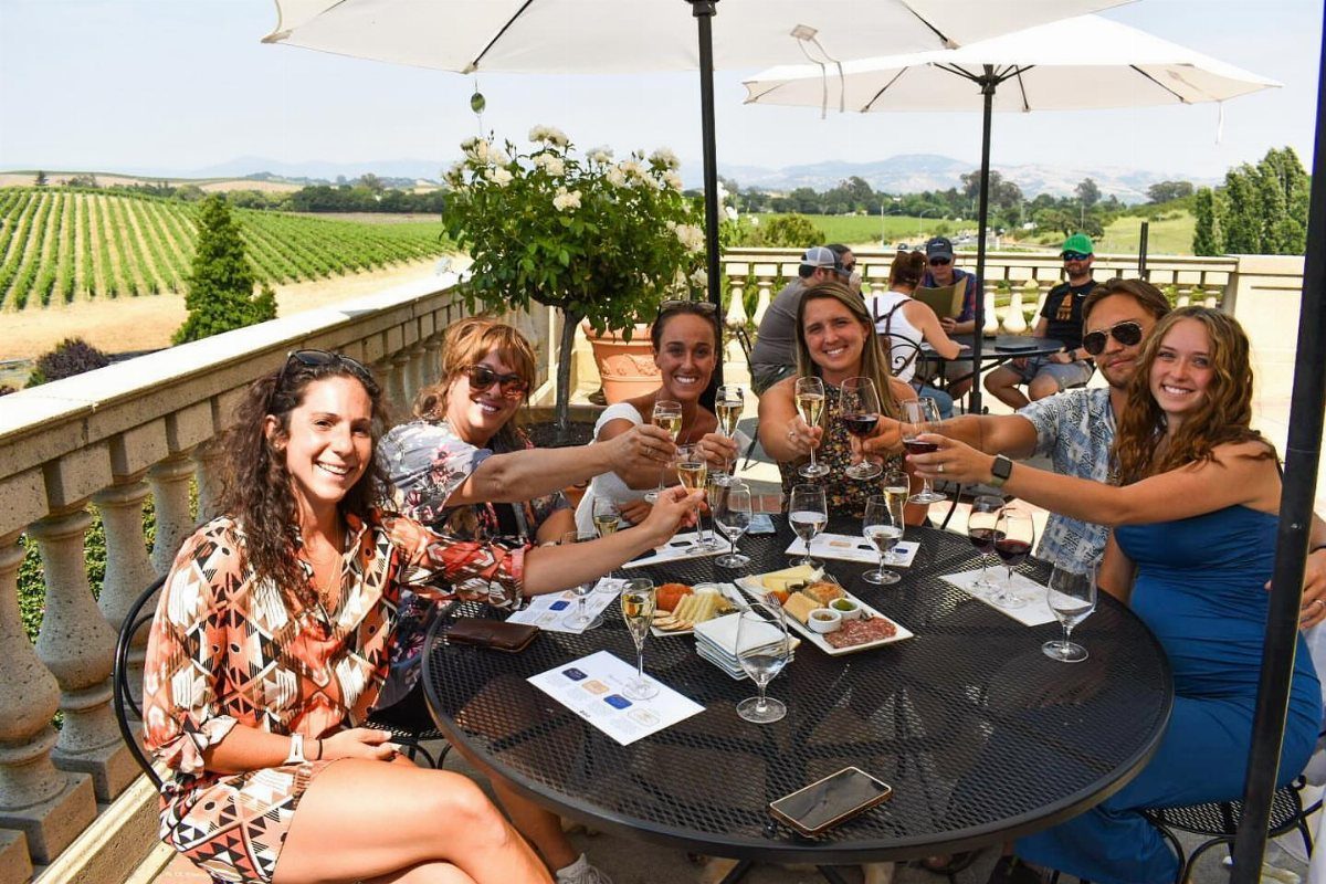 Women clinking wine glasses on a terrace overlooking lush green vineyards and mountains.