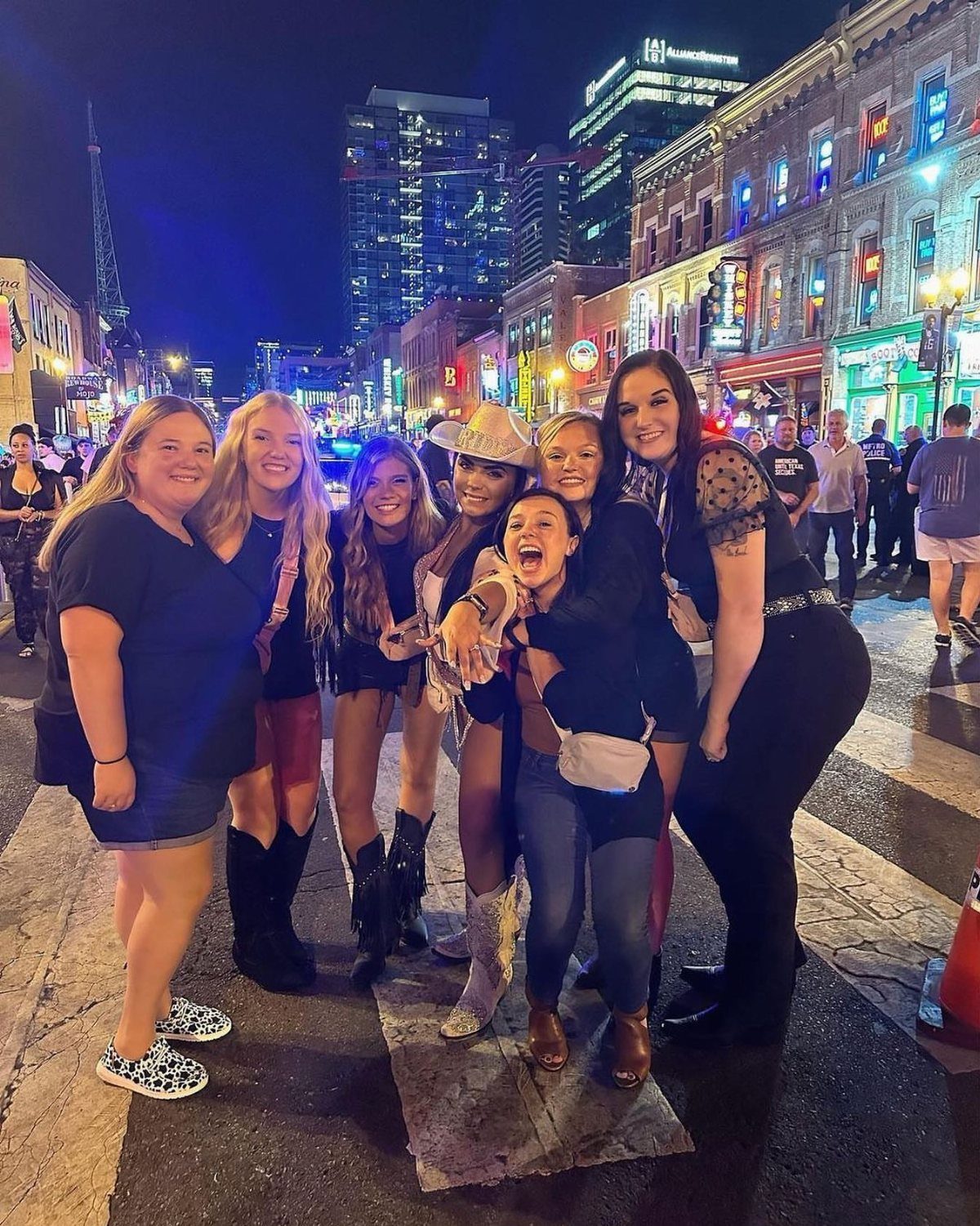 Six young women in cowboy boots posing on a neon-lit city street at night.
