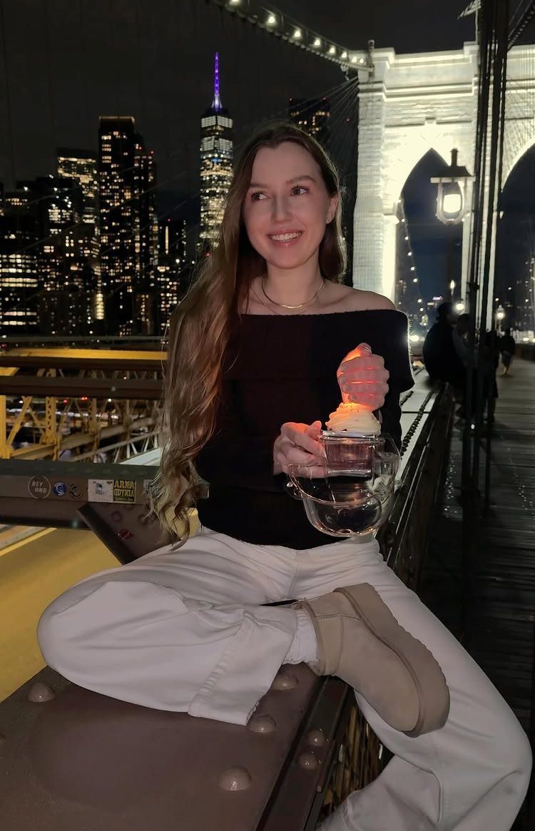 Young woman sitting on a bridge railing with New York City skyline at night