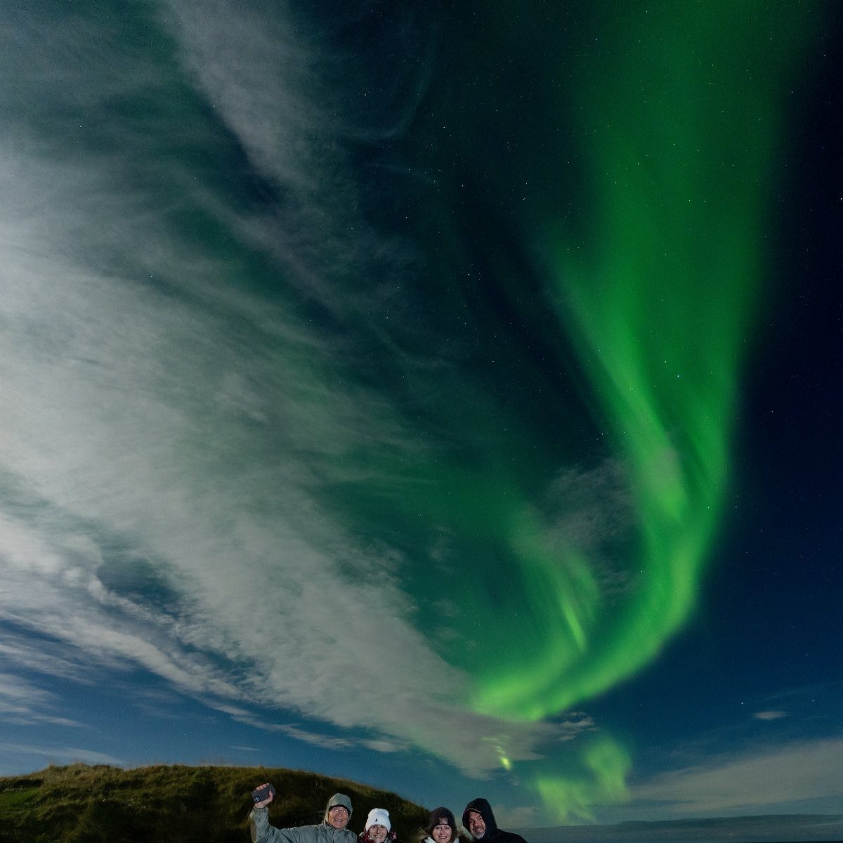 Four people standing under a bright green aurora borealis in a night sky.