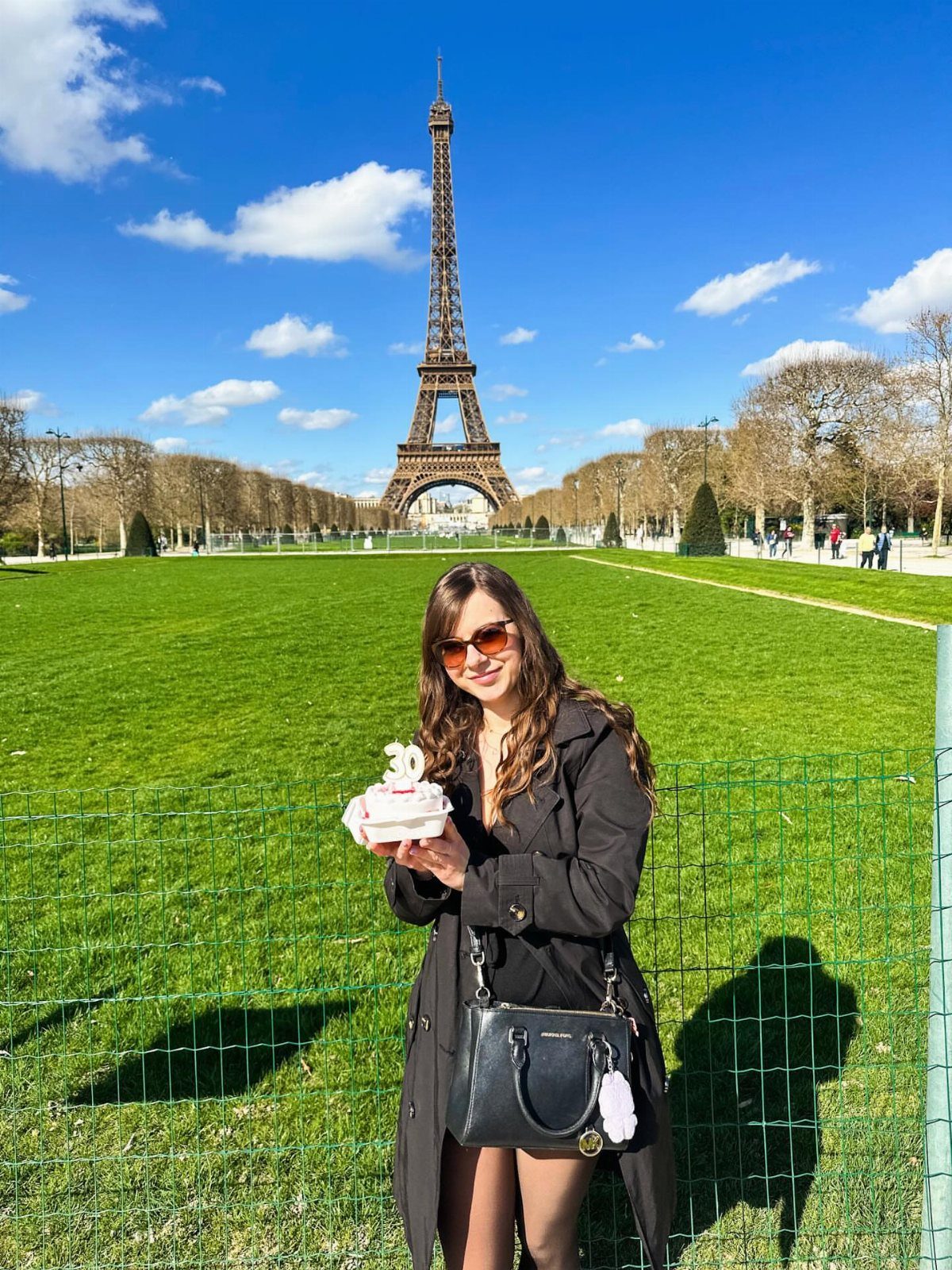 Woman holding a birthday cake in front of the Eiffel Tower in Paris.
