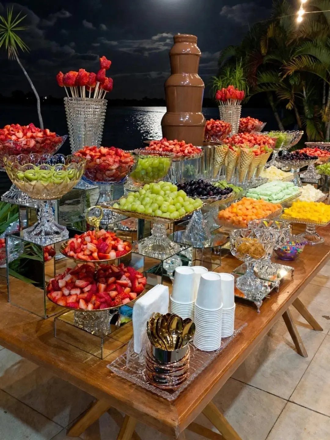 Lavish dessert table with chocolate fountain and fresh fruit bowls outdoors at night.