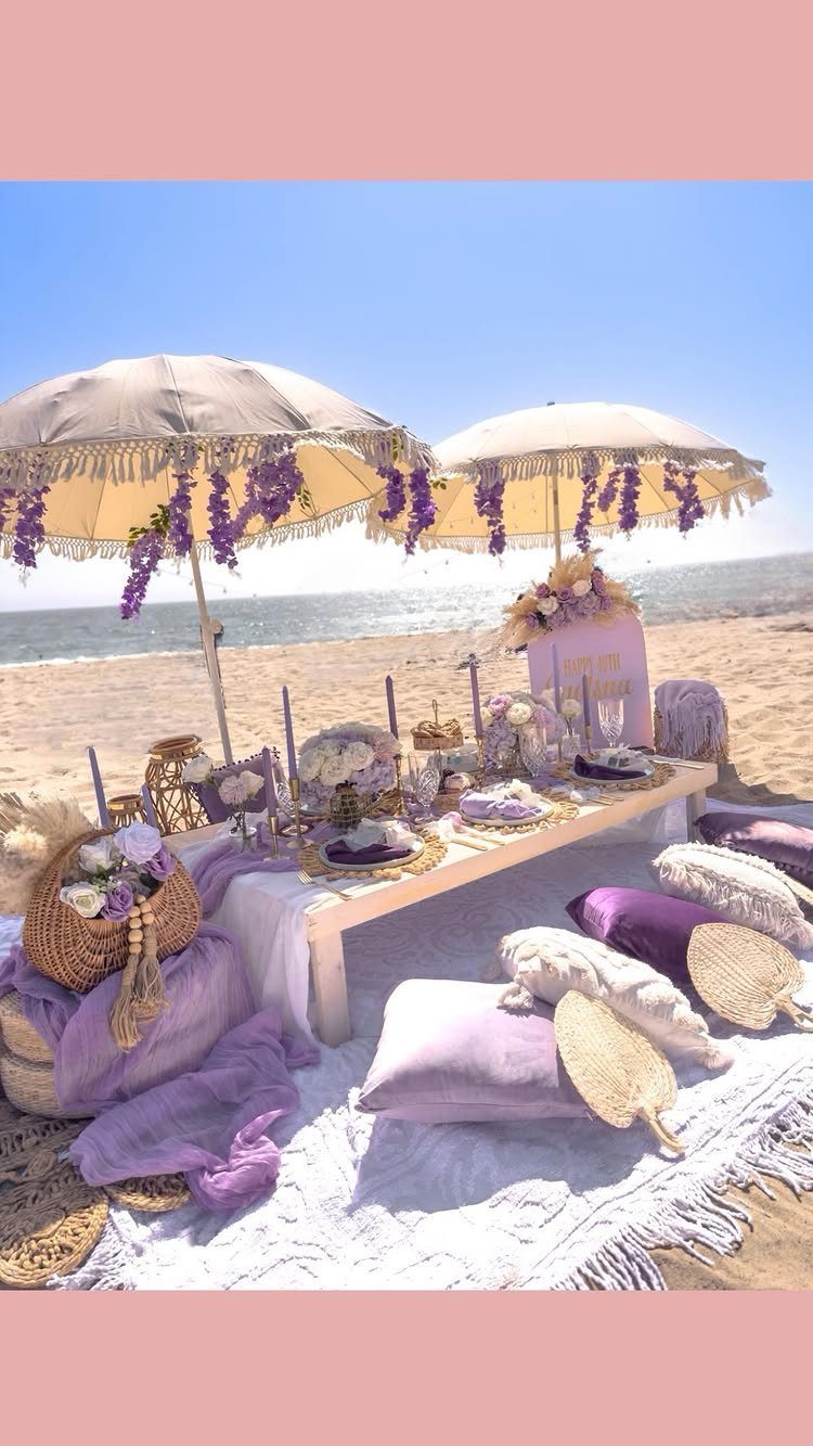 Luxurious beach picnic setup with purple cushions and umbrellas on a sandy shore.