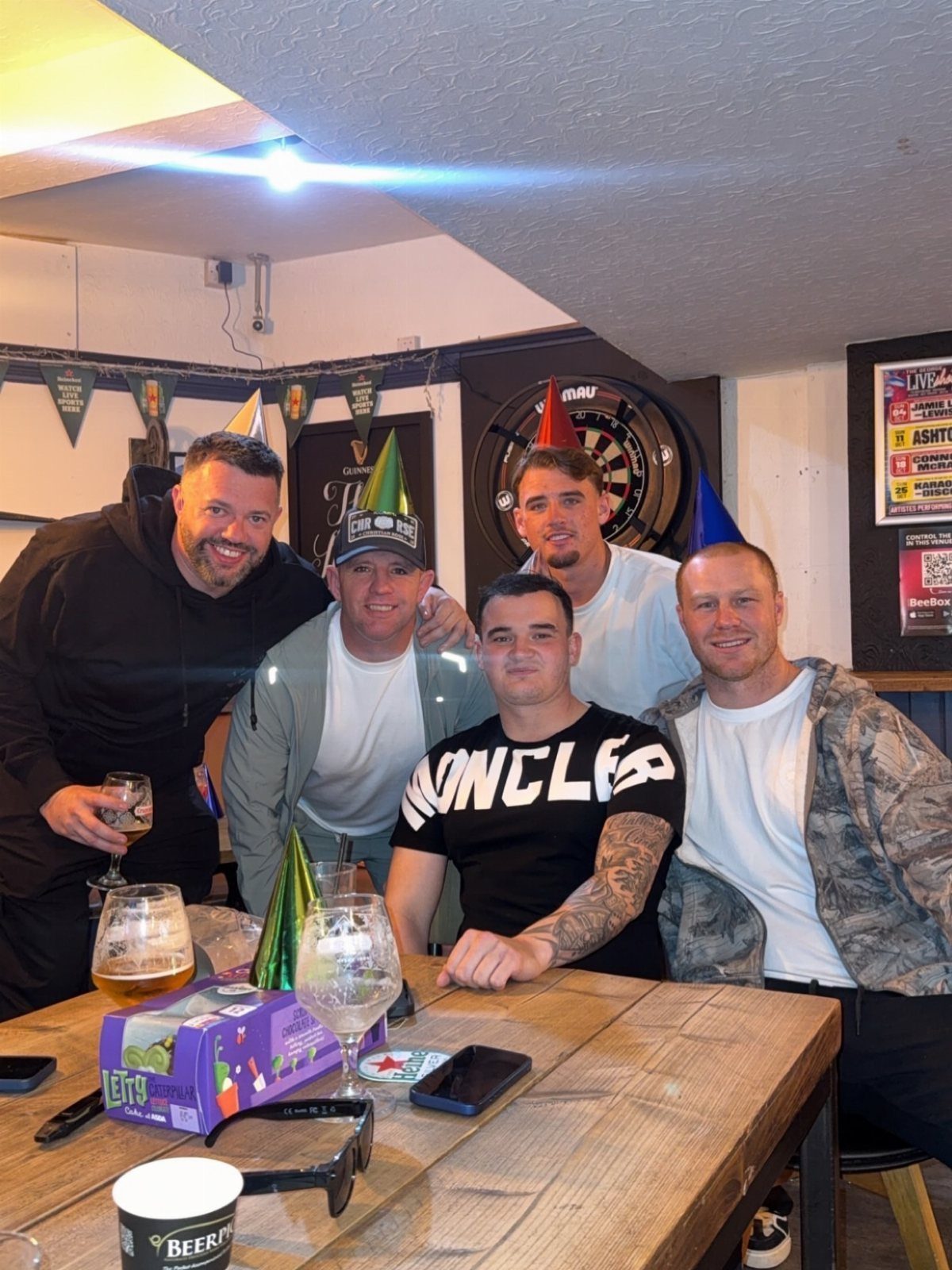Five men wearing party hats posing around a table with beer in a pub.