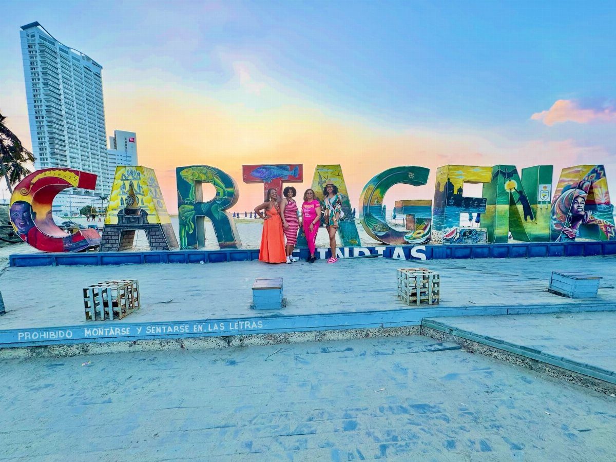 Three women posing before large colorful Cartagena letters on a sunny waterfront promenade.