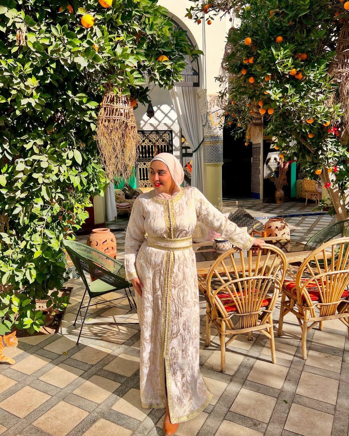 Woman in traditional Moroccan kaftan standing in a sunny courtyard with orange trees.