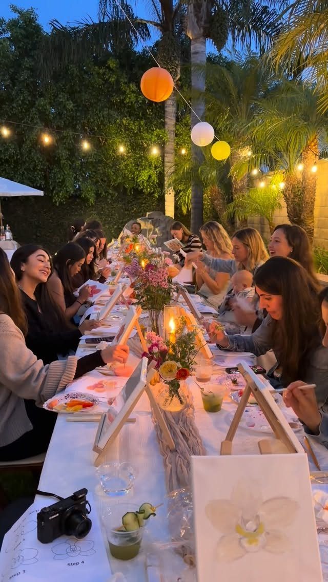 Women painting on small canvases at a decorated outdoor dinner party at dusk
