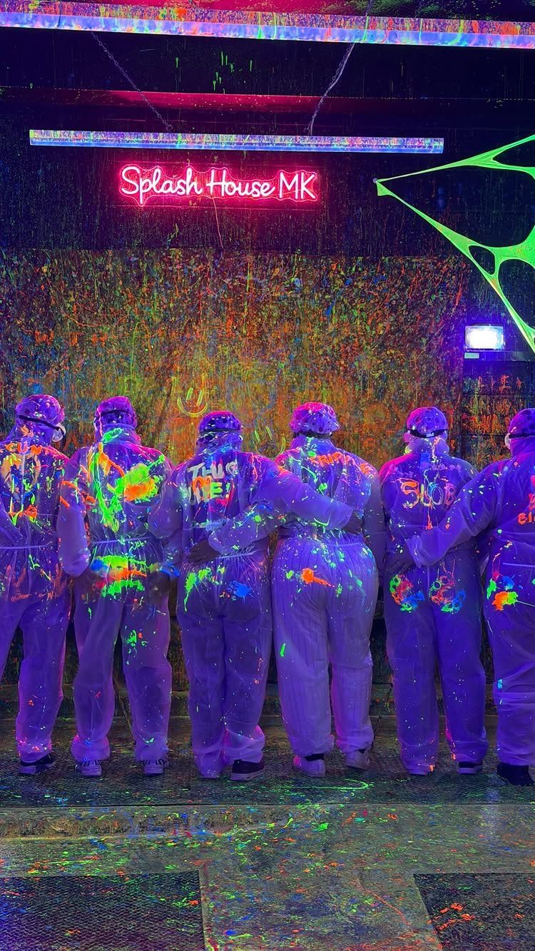 Six friends in paint-splattered white suits posing under a neon Splash House sign.