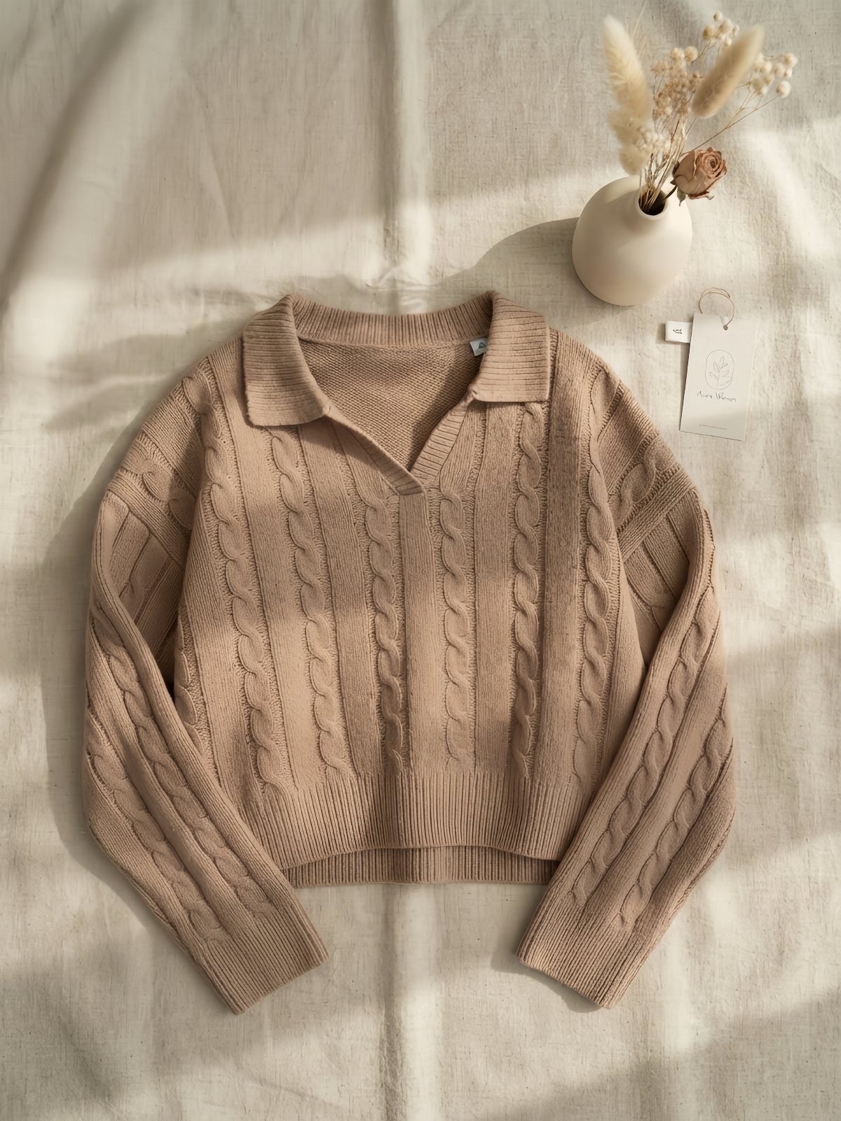 Beige cable-knit sweater on linen fabric with a small vase of dried flowers