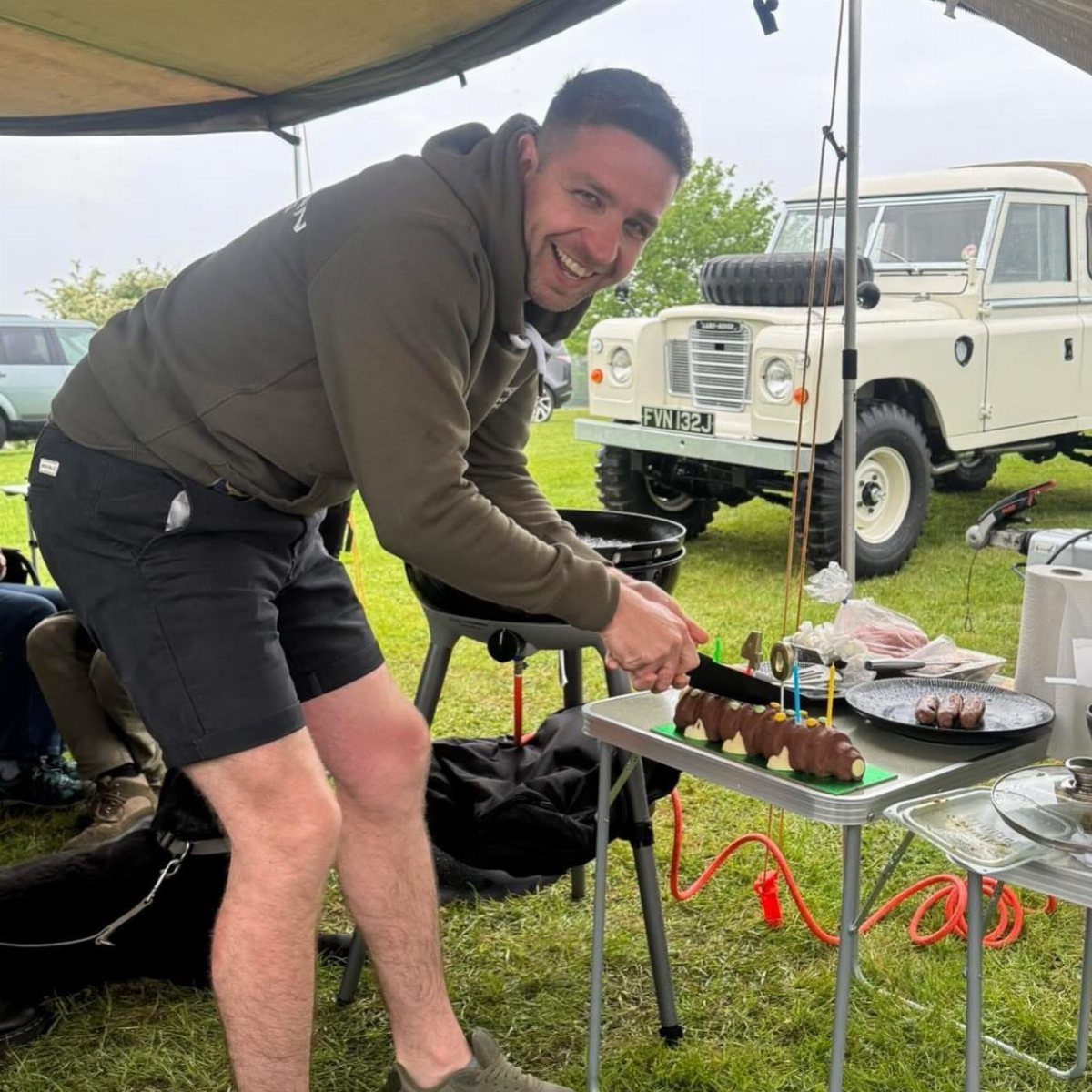 Man grilling meat under a canopy with a vintage white off-road vehicle behind him.