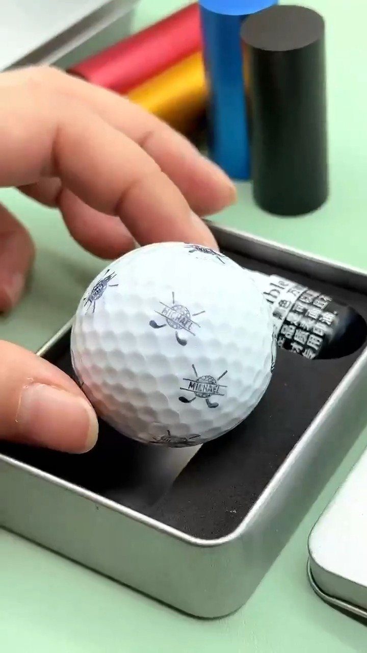 White golf ball with printed logos held over a silver metal gift tin.