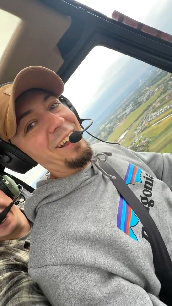Smiling man in a headset sitting inside a helicopter overlooking a coastal landscape.