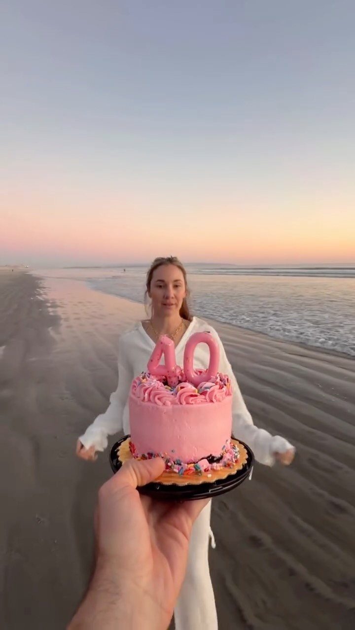 Pink birthday cake with 40 candle held in front of woman on beach