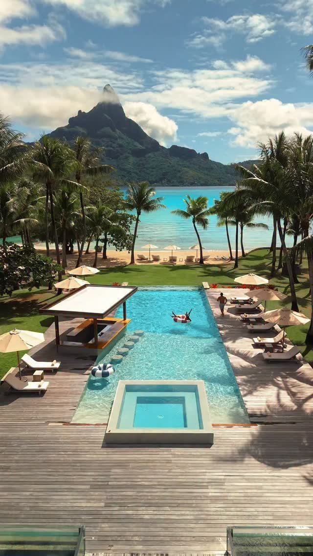 Luxury resort pool and wooden deck overlooking a tropical beach and mountain peak.