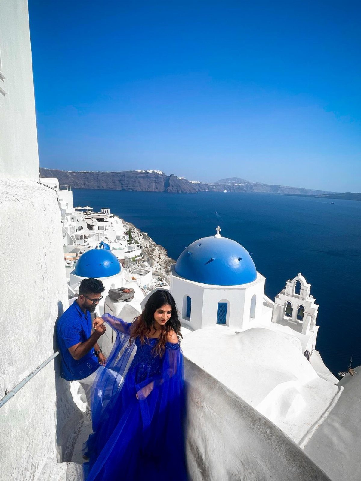 Couple in blue attire posing against white buildings and blue domes in Santorini