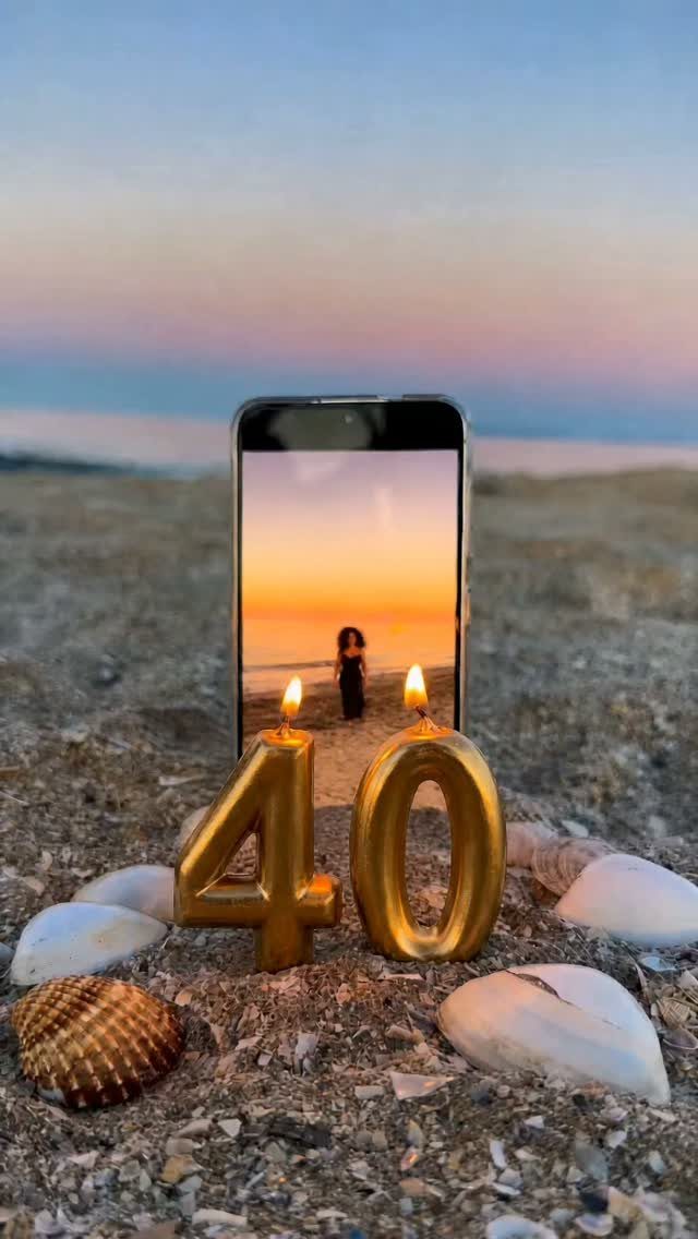 Gold number 40 candles lit on a beach with a smartphone displaying a sunset.