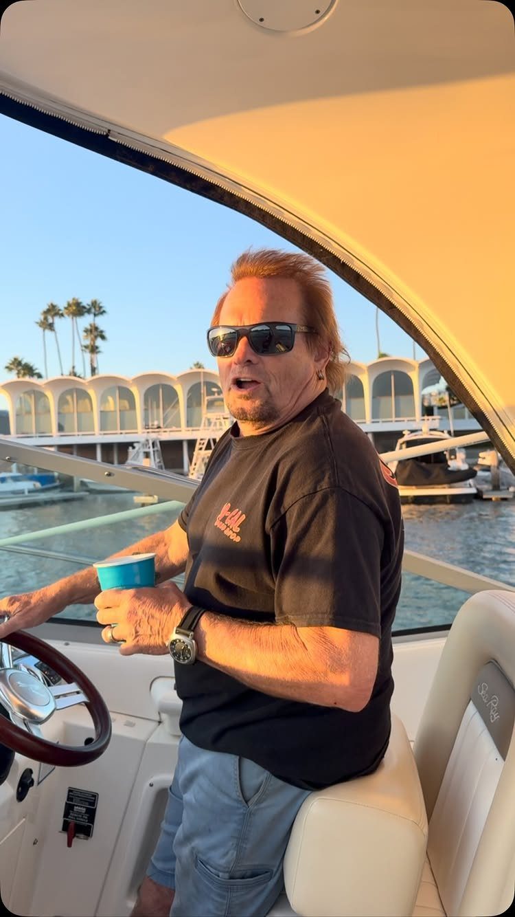 Middle-aged man with sunglasses sitting at a boat helm in a sunny marina.