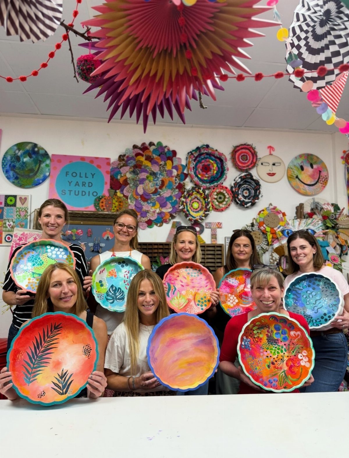 Eight women holding colorful hand-painted ceramic plates in a bright art studio.