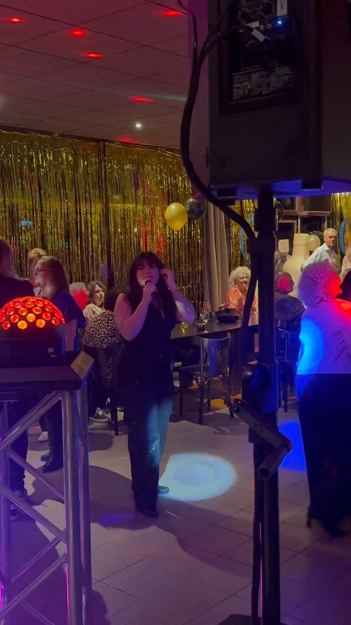 Woman singing into a microphone at a party with gold tinsel backdrop.