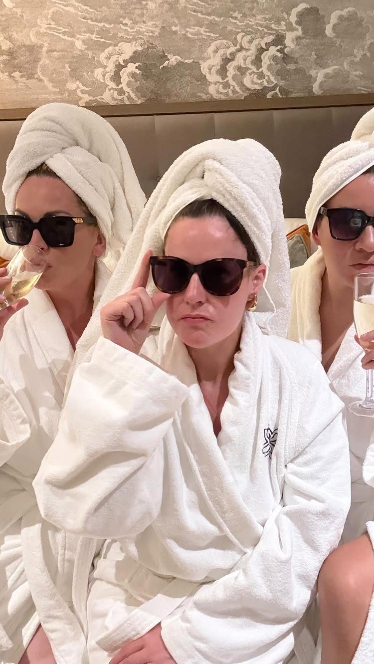 Three women in white robes and towel head wraps wearing sunglasses and drinking champagne.