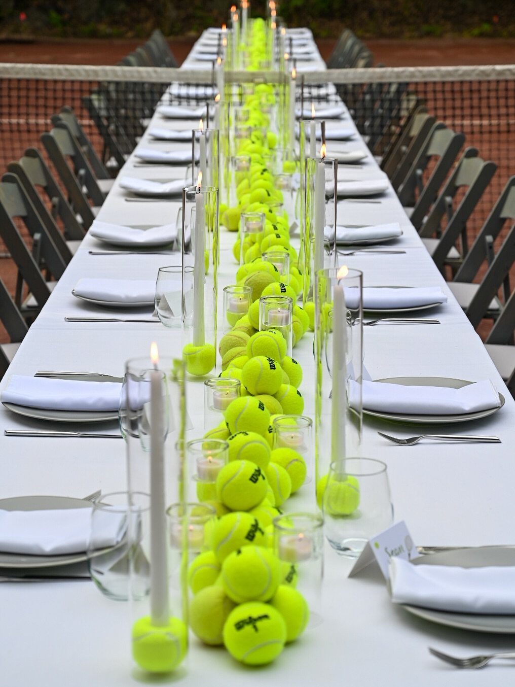 Long white table on a tennis court decorated with yellow tennis balls and candles.