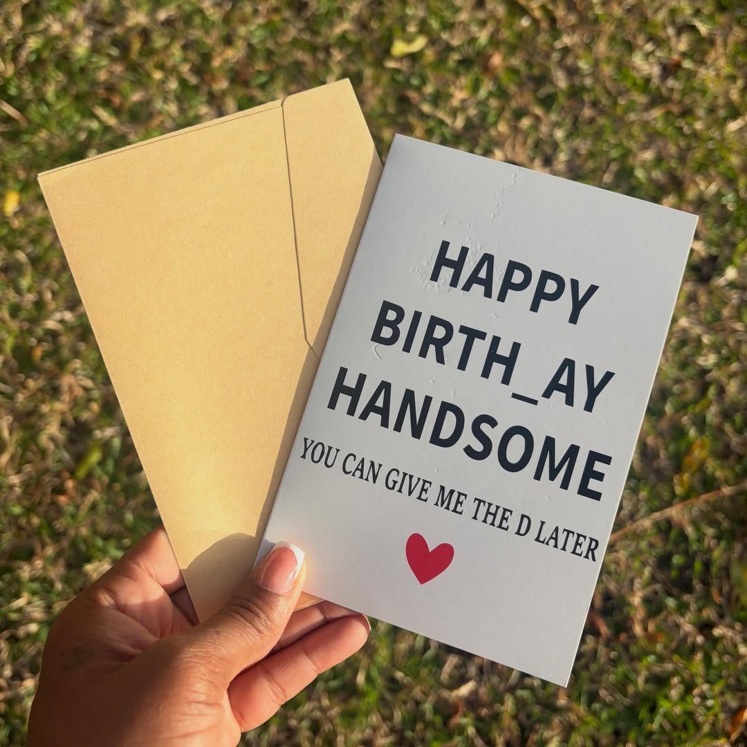 White greeting card with cheeky text and red heart held over grass